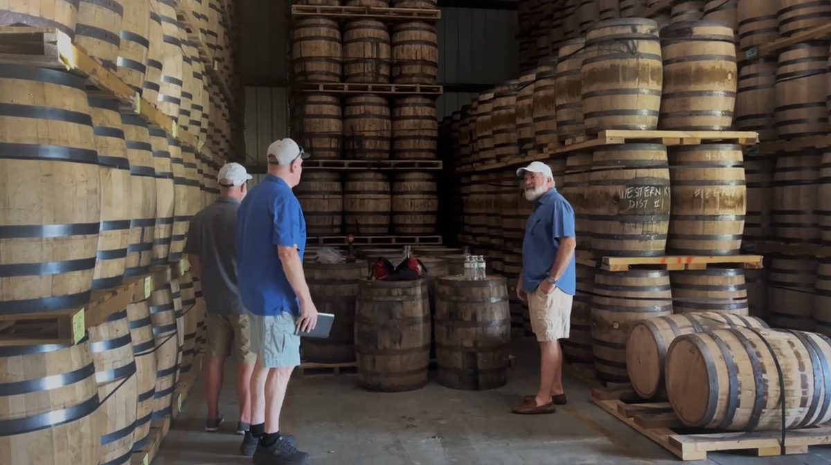 Three men standing among stacked barrels inside a rickhouse
